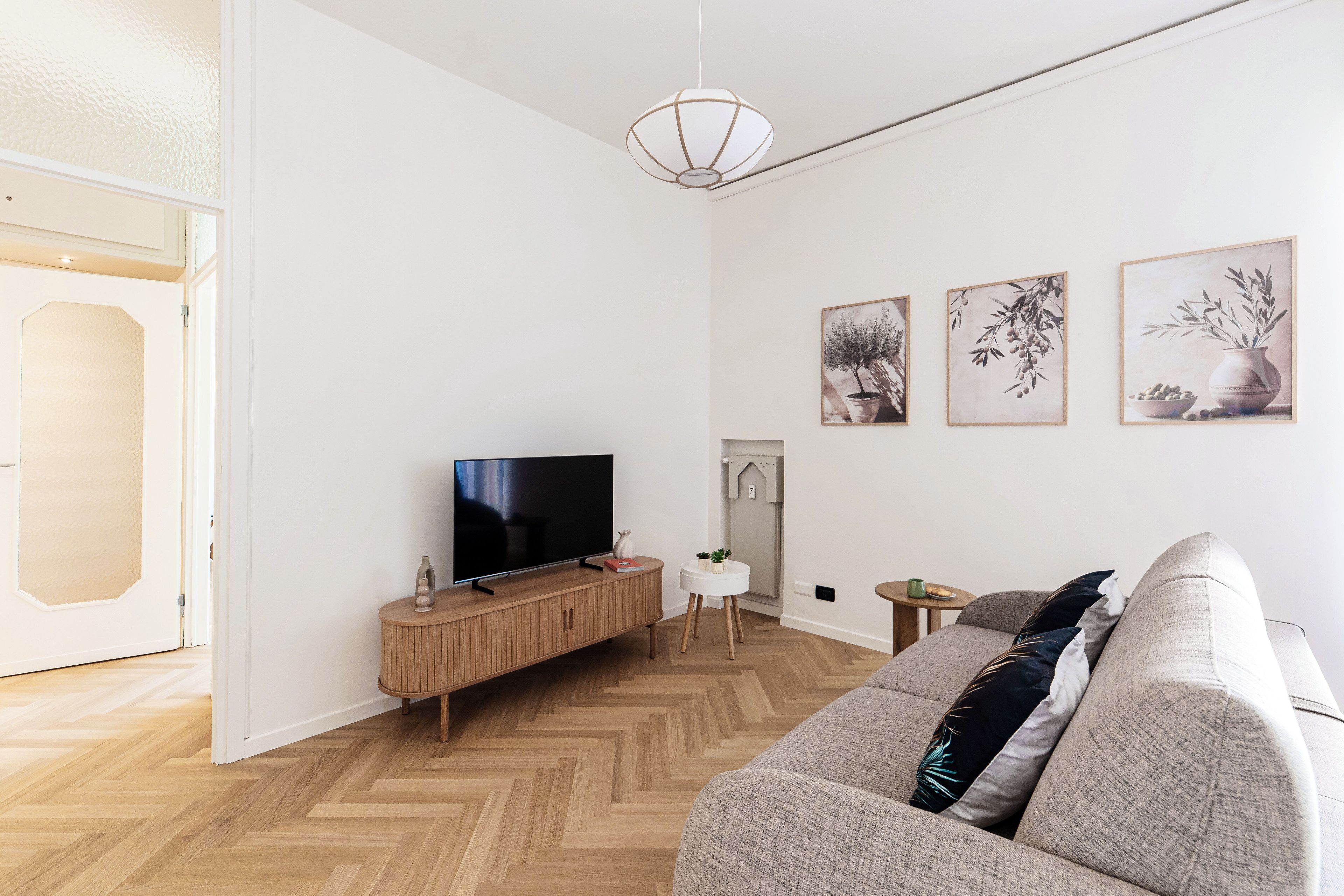 A modern living room with a flat-screen TV on a wooden console, a grey sofa, and three framed botanical prints on the wall.