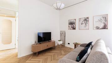A modern living room with a flat-screen TV on a wooden console, a grey sofa, and three framed botanical prints on the wall.