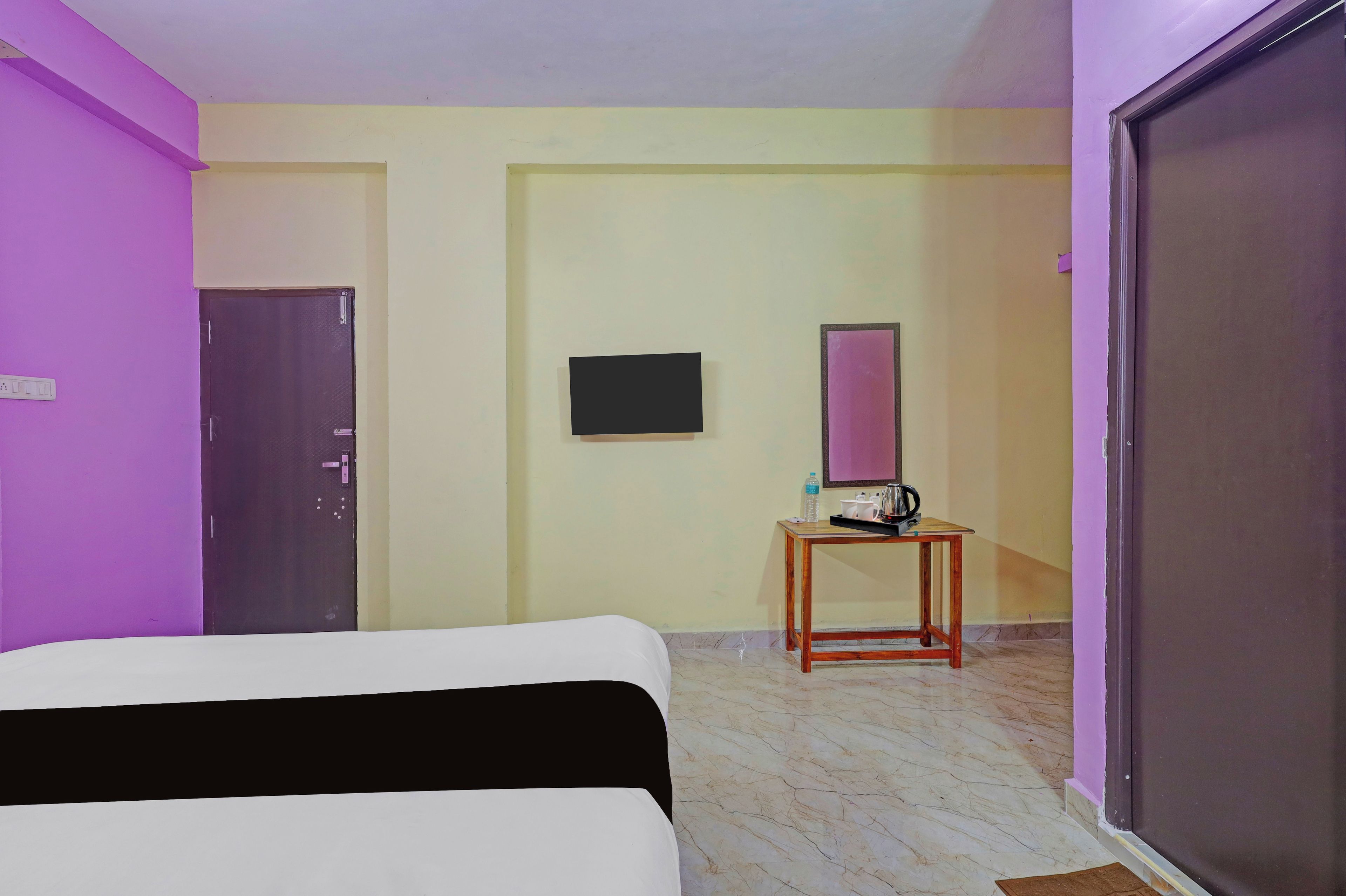 Habitación de hotel con una cama, una mesita con hervidor de agua, una botella de agua y un televisor en la pared.