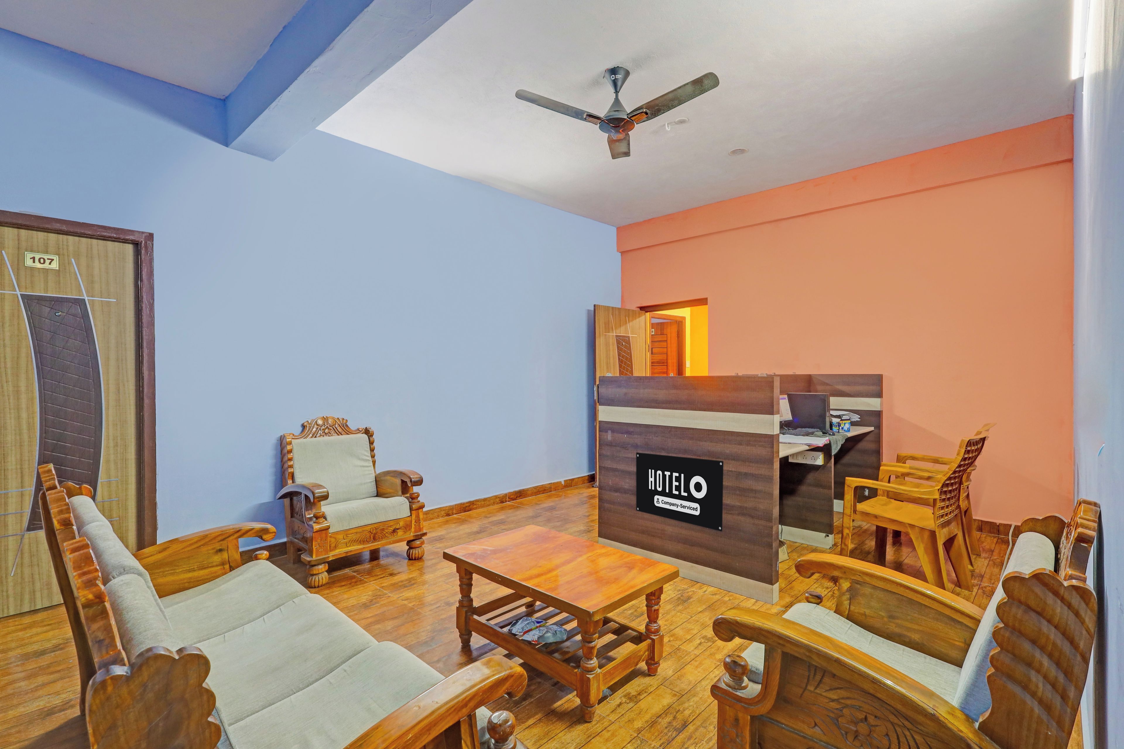 A hotel room with a wooden sofa set, a coffee table, a ceiling fan, and a sign indicating the room is company serviced.
