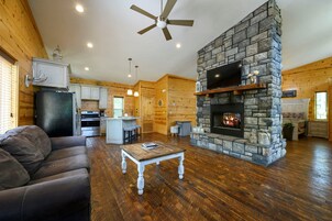 A log cabin interior with a stone fireplace, wooden floors, a ceiling fan, and a kitchen area.