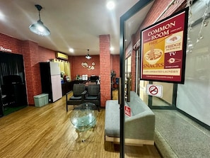A common room with a brick wall, a sofa, a glass coffee table, and a sign advertising snacks and coin-operated laundry. - The Lin Fifty (Klong Toei)