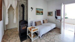 Uma sala de estar acolhedora com piso estampado em pedra, um sofá branco com almofadas bege, uma mesinha com objetos decorativos, uma lareira a lenha de madeira preta e uma janela com vista para o verde.