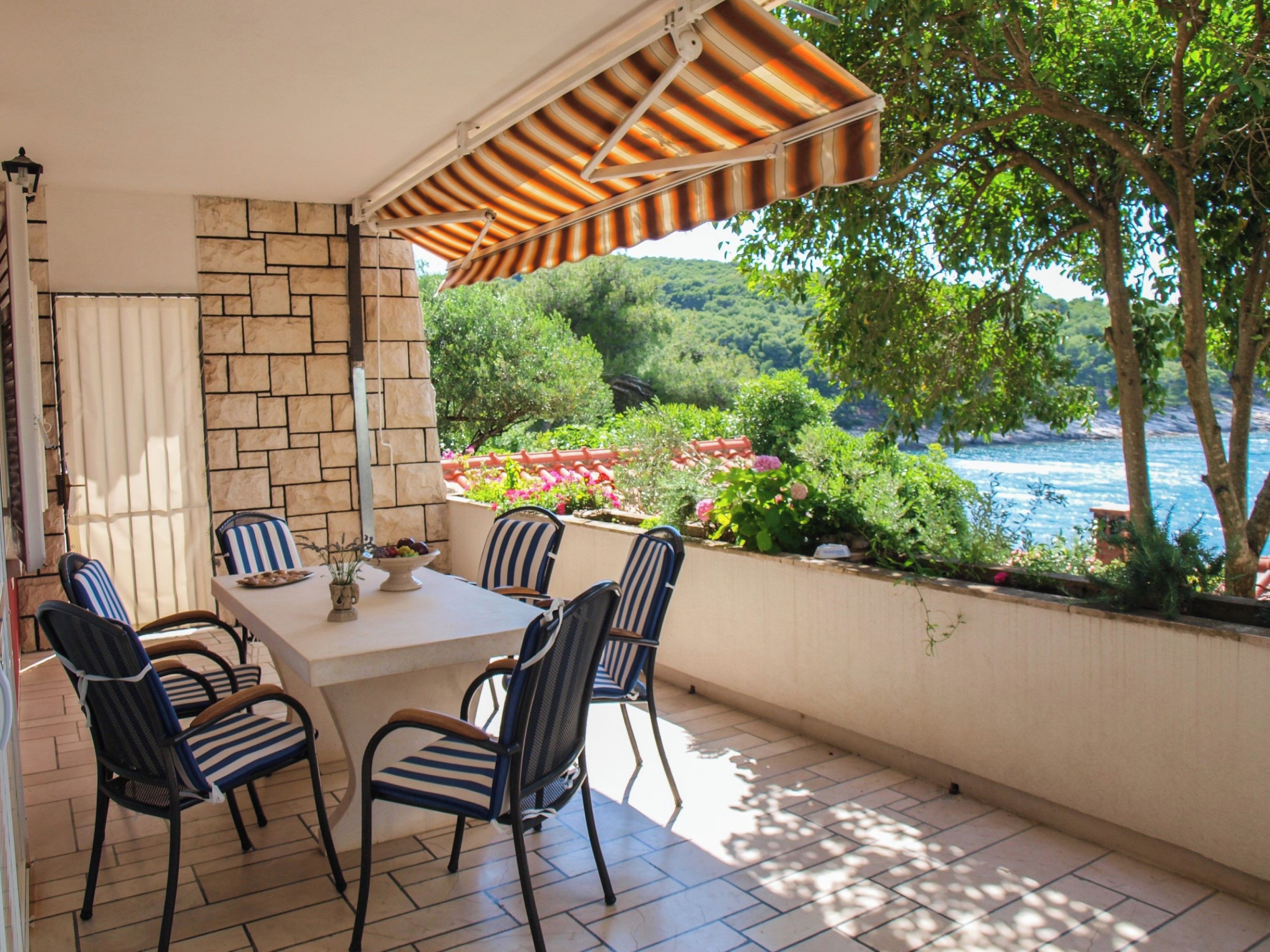 Une terrasse extérieure avec une table et des chaises, un mur en pierre et une vue sur une étendue d’eau et de la verdure.
