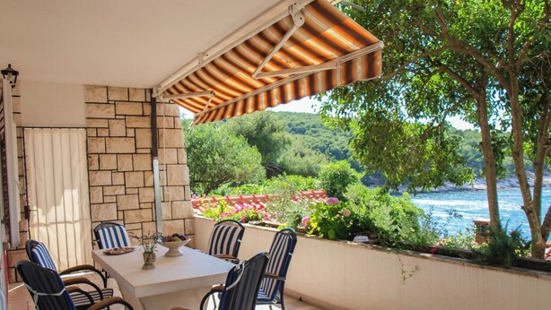 Um pátio externo com mesa e cadeiras, uma parede de pedra e vista para a água e vegetação.