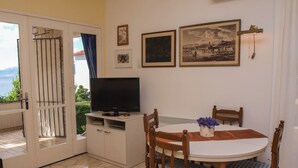 Uma sala de jantar com mesa, cadeiras, uma televisão e vista para o mar através de uma porta de vidro.