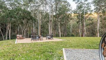 Un espace extérieur aménagé avec deux chaises et une table, entouré d’un champ herbeux et de grands arbres.