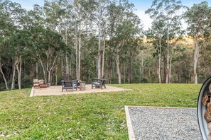 An outdoor seating area with two chairs and a table, surrounded by a grassy field and tall trees.