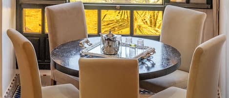 A round table with four chairs, a decorative tray, and a view of greenery through large windows.