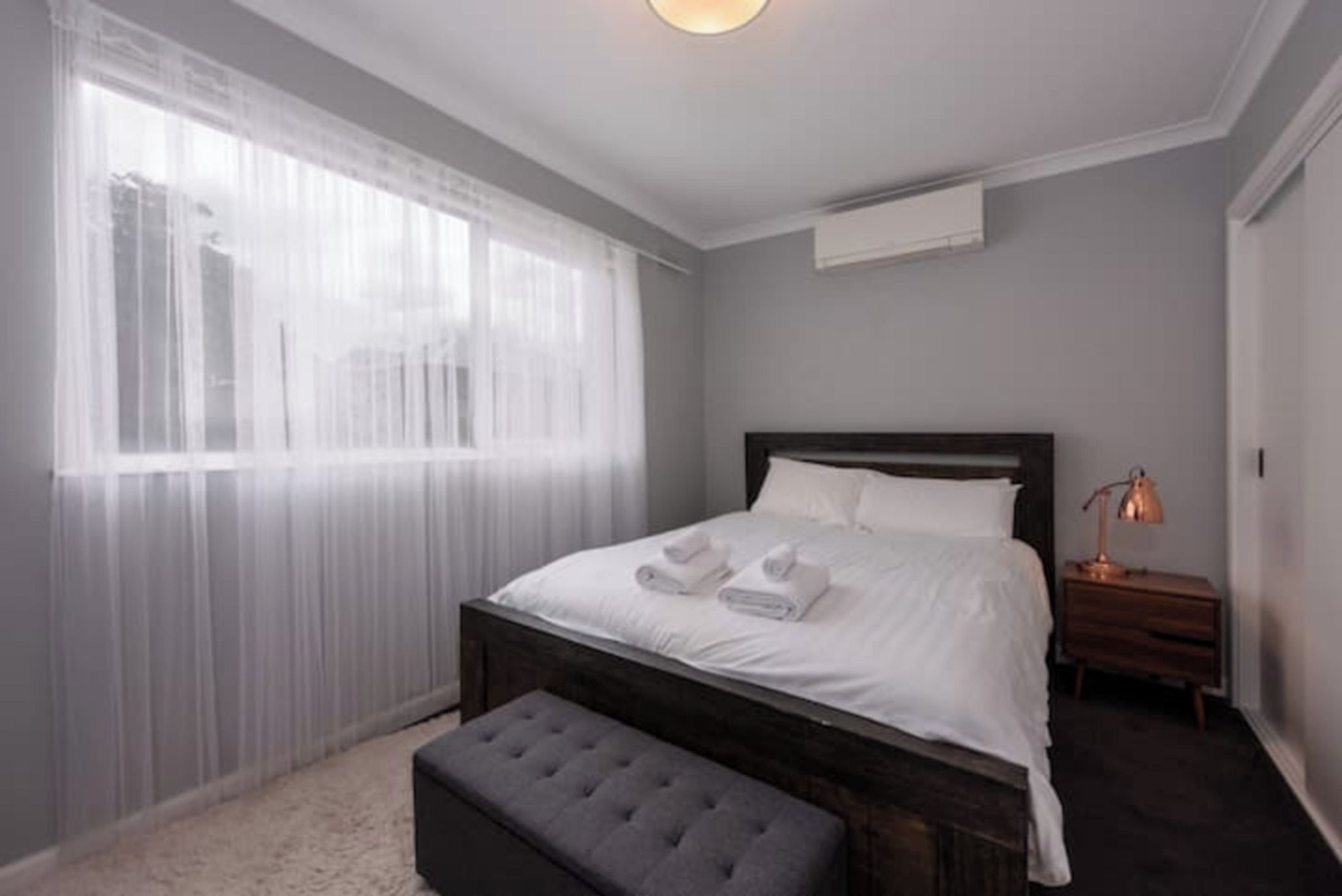 A bedroom with a bed, a bedside table, a lamp, and a window with sheer curtains.