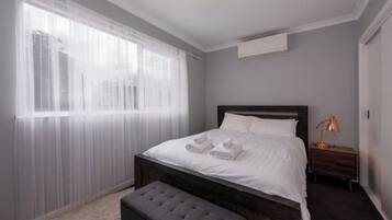 A bedroom with a bed, a bedside table, a lamp, and a window with sheer curtains.
