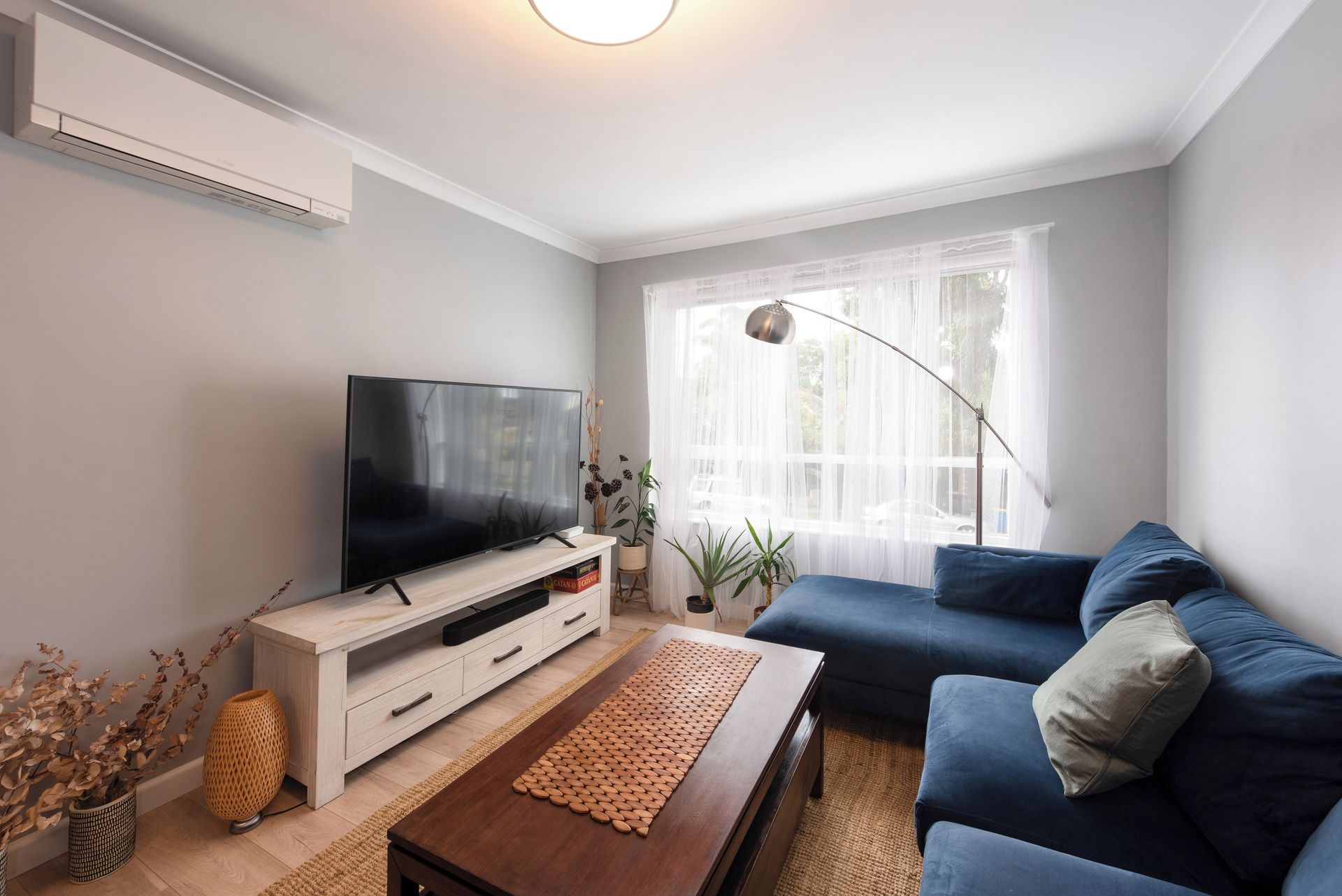 A living room with a flat-screen TV, a blue sofa, a wooden coffee table, and a large window with sheer curtains.