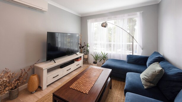 Una sala de estar con un televisor de pantalla plana, un sofá azul, una mesa de centro de madera y un ventanal con cortinas translúcidas.
