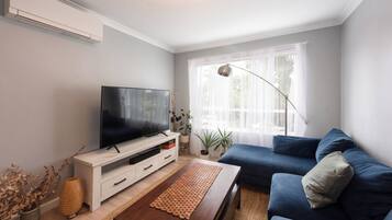 A living room with a flat-screen TV, a blue sofa, a wooden coffee table, and a large window with sheer curtains.