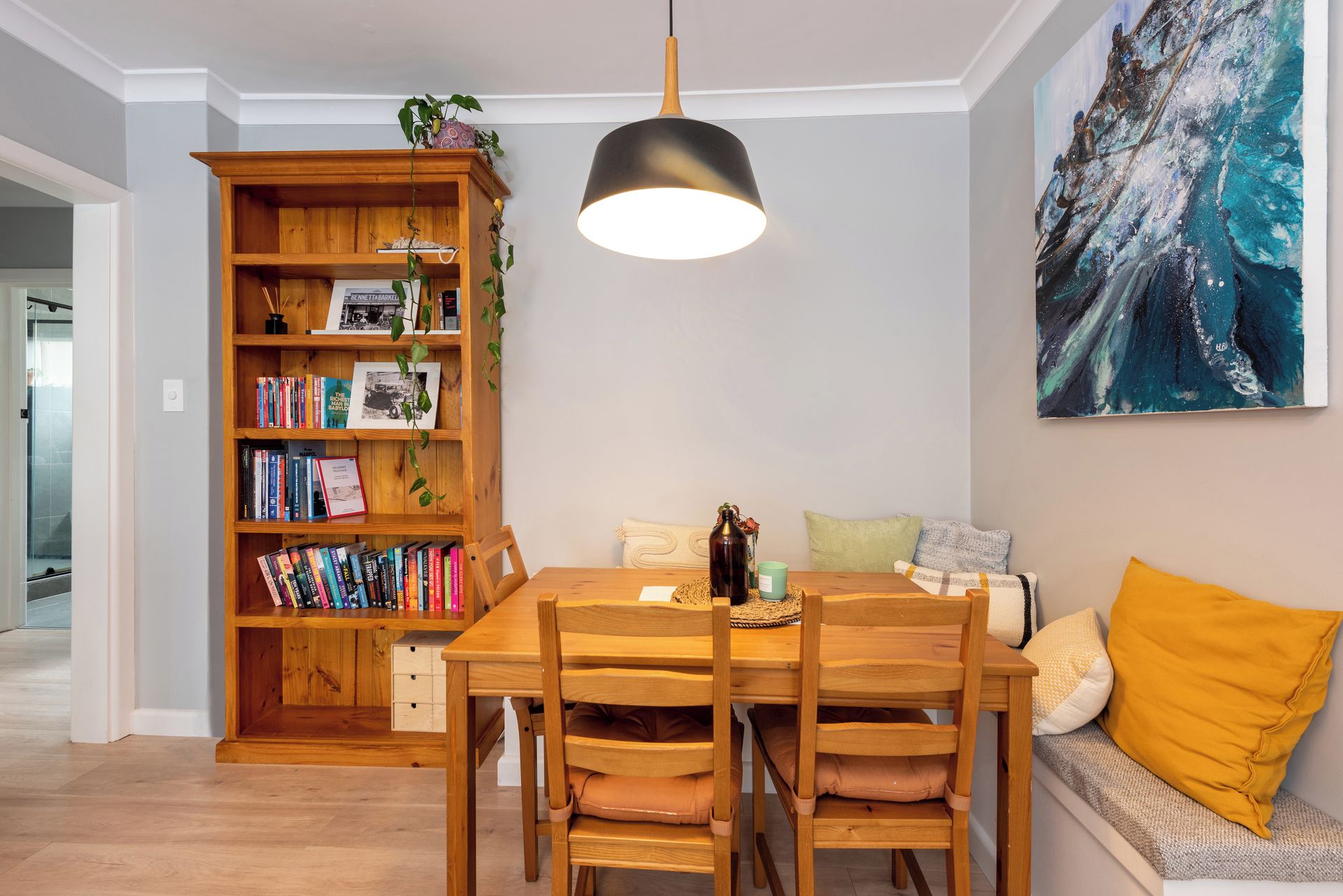A dining area with a wooden table and chairs, a bookshelf, a hanging pendant light, and a painting on the wall.
