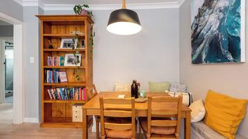 A dining area with a wooden table and chairs, a bookshelf, a hanging pendant light, and a painting on the wall.