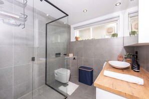 A modern bathroom with a glass shower enclosure, a toilet, a sink with a wooden countertop, and a window with plants. - Homely and Cozy 1BDR Unit in Leafy Hawthorn (Hawthorn)