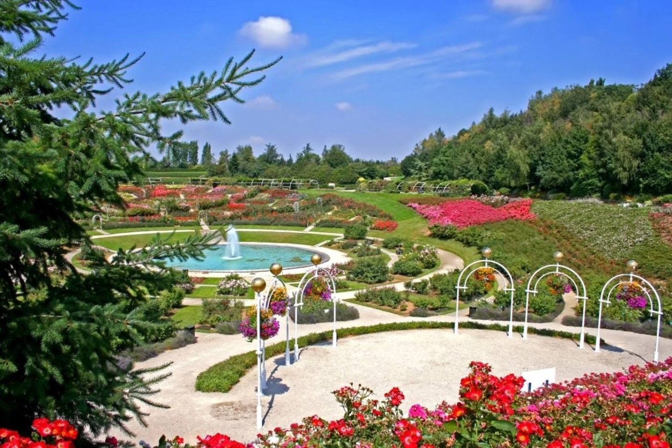 Un jardin aménagé avec une fontaine, des parterres de fleurs et un ciel dégagé.