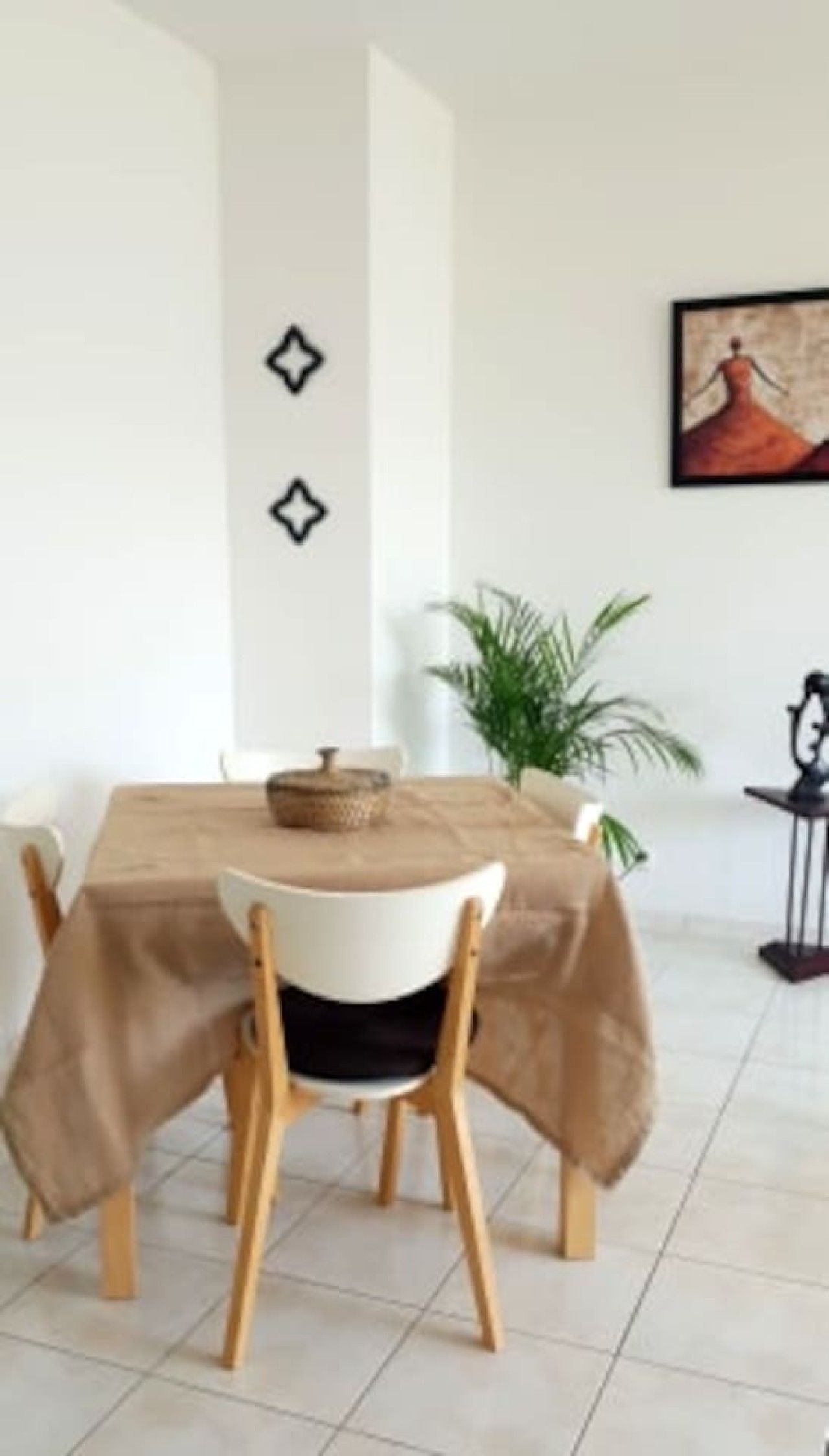 Ein Essbereich mit einem Tisch, der mit einer beigen Tischdecke bedeckt ist, Holzstühle, einer Zimmerpflanze und einem gerahmten Kunstwerk an der Wand.