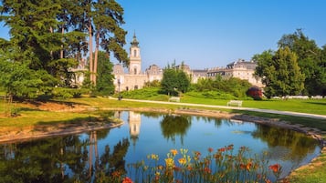 A large building with a prominent tower, surrounded by trees and a calm water body with flowers in the foreground.