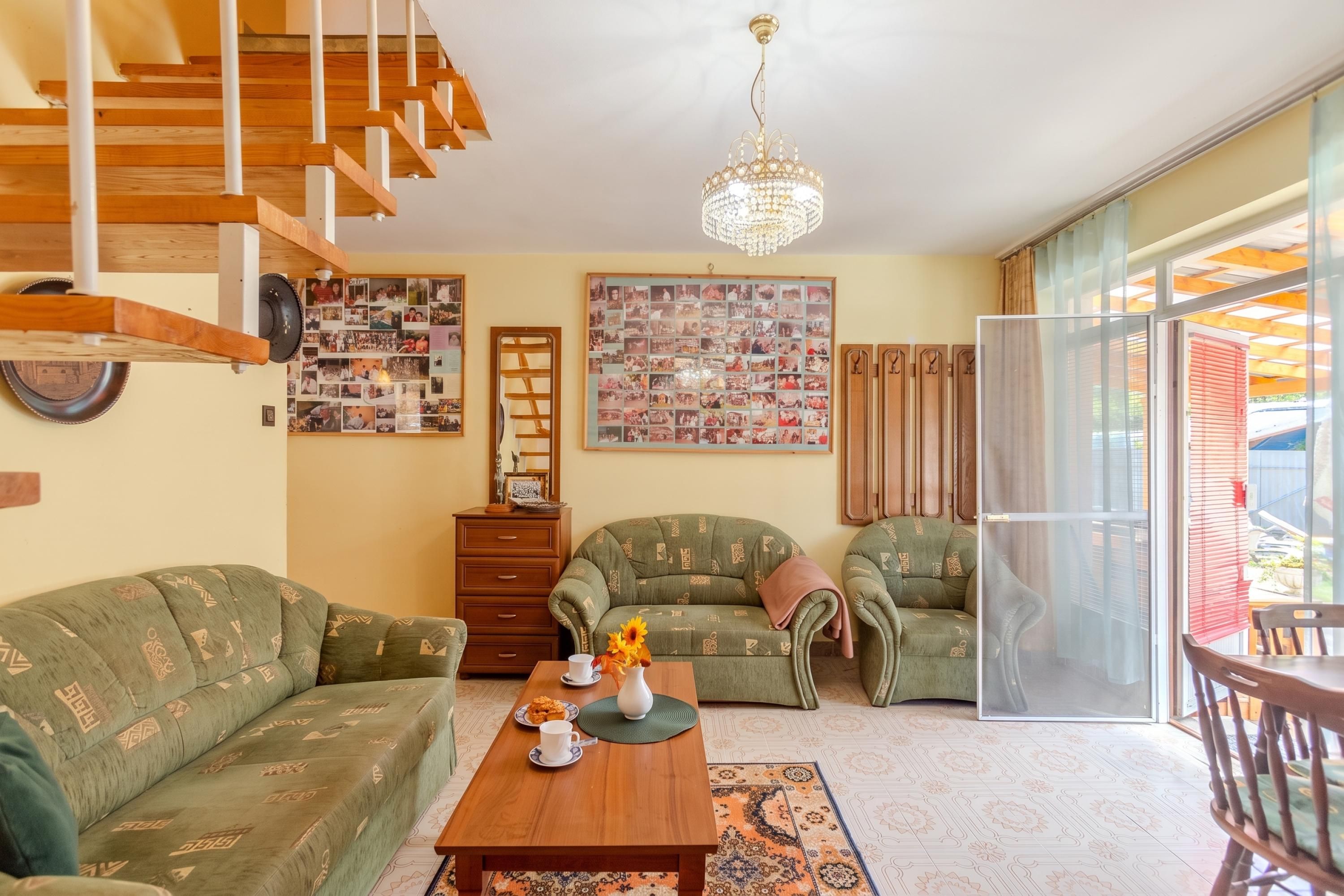 A living room with a wooden staircase, a chandelier, two green patterned sofas, a coffee table with a vase and a plate of cookies, and a large photo collage on the wall.