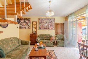 Una sala de estar con una escalera de madera, un candelabro, dos sofás con estampado verde, una mesa de centro con un jarrón y un plato de galletas, y una gran composición de fotos en la pared.