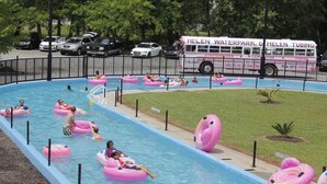 Une piscine avec des personnes sur des bouées gonflables, un autobus arborant la marque « Helen Waterpark & Helen Tubing », et un espace clos entouré d’arbres.