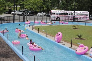 A swimming pool with people on inflatable rings, a bus with 'Helen Waterpark & Helen Tubing' branding, and a fenced area with trees. - 🌷 Das Tulip of Helen. Center of town, Ground floo (Helen)