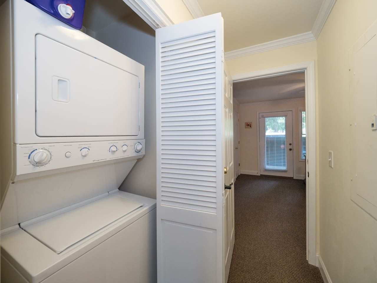 A laundry room with a washer and dryer, a door leading to a hallway, and a carpeted floor.