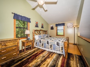 A bedroom with a wooden bed, a desk, and a chair. There is a ceiling fan, a window with blinds, and a lamp.