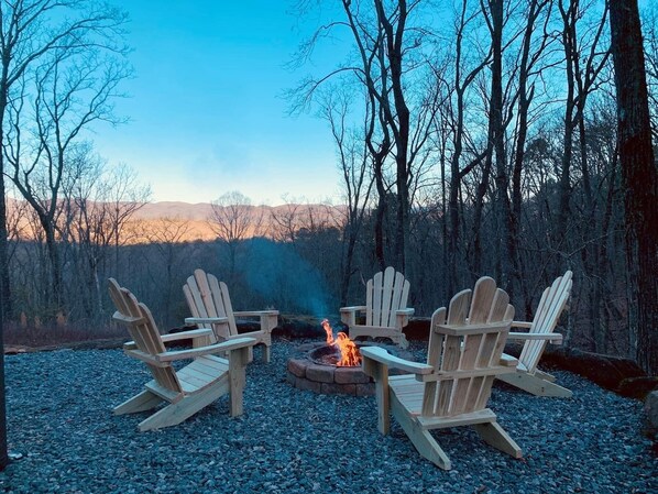 A fire pit with four Adirondack chairs arranged around it, set on a gravel surface with trees in the background.
