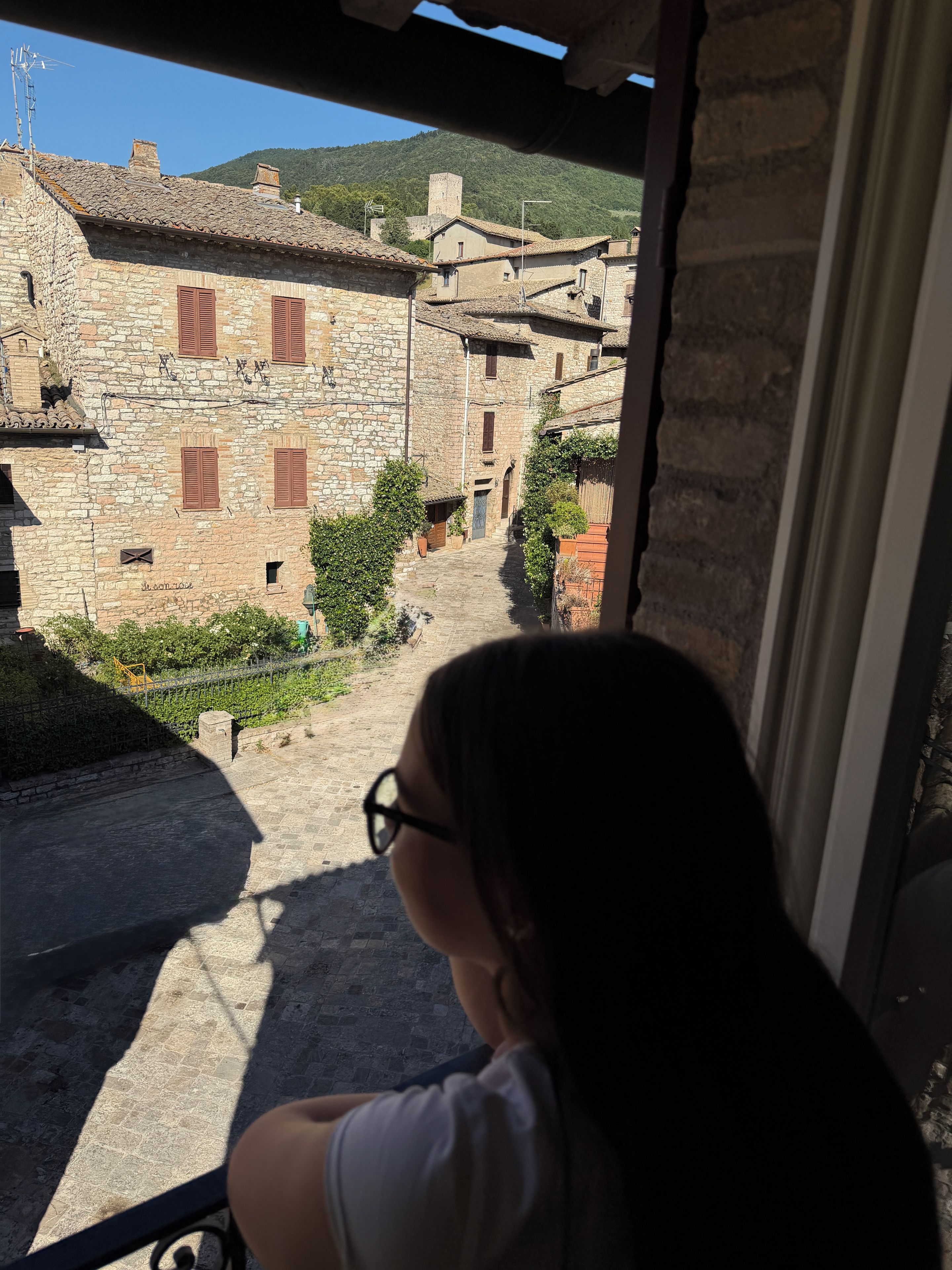 Un edificio in pietra con una stretta via che vi conduce, cielo sereno e una persona seduta su un balcone.