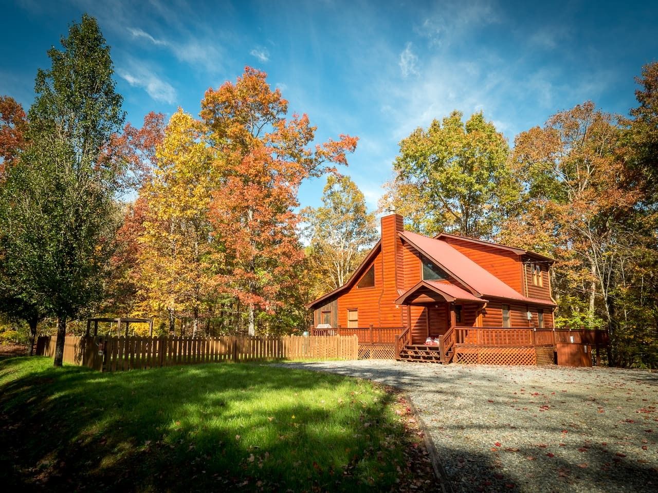 A wooden cabin with a sloped roof, surrounded by a wooden fence and trees with autumn foliage.