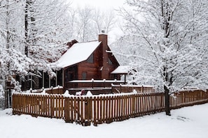 Una cabaña de troncos con techo nevado y valla de madera alrededor.