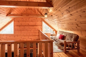 Un intérieur en bois avec une chambre à coucher située en mezzanine, une balustrade en bois et un canapé agrémenté d’un coussin rouge.