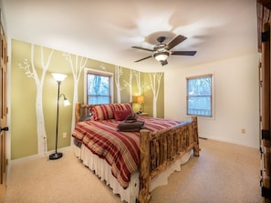 Une chambre avec un lit en bois, un ventilateur de plafond et deux fenêtres équipées de stores.