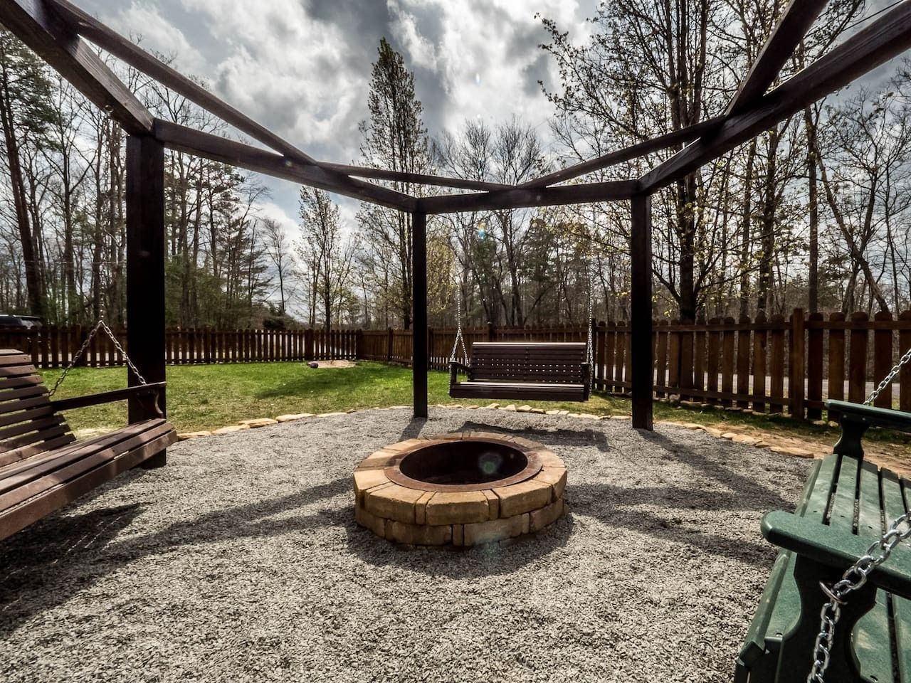 A wooden swing set with a circular seating area in the middle, surrounded by a gravel ground.