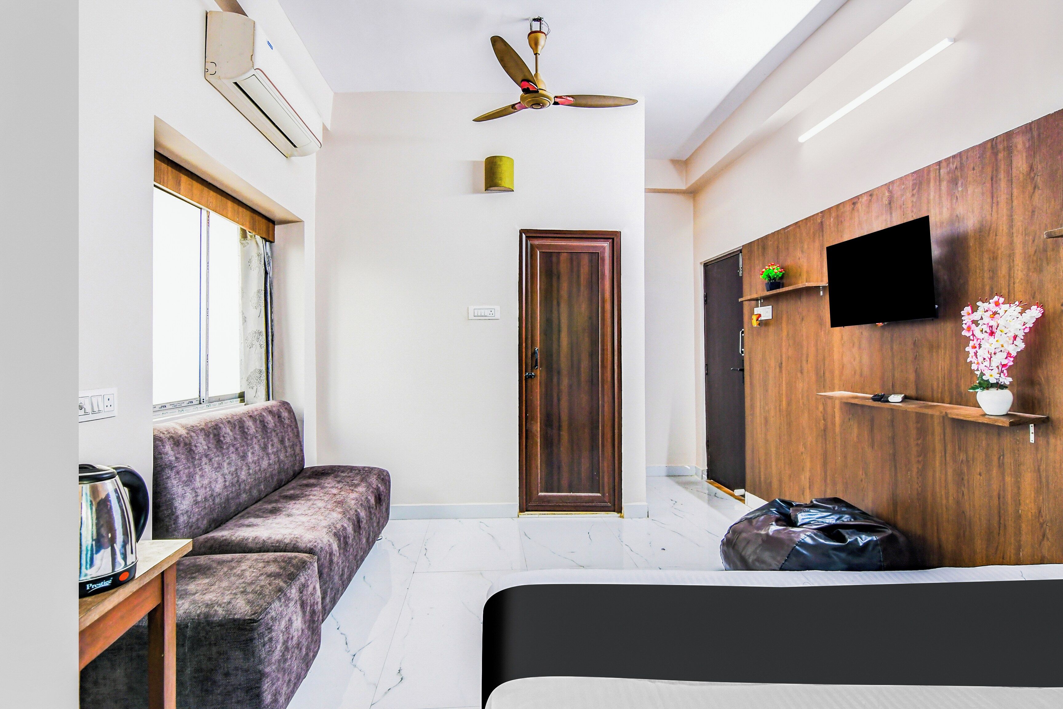 Quarto de hotel moderno com sofá, cama, ventilador de teto, TV presa a uma parede de madeira e uma janela.