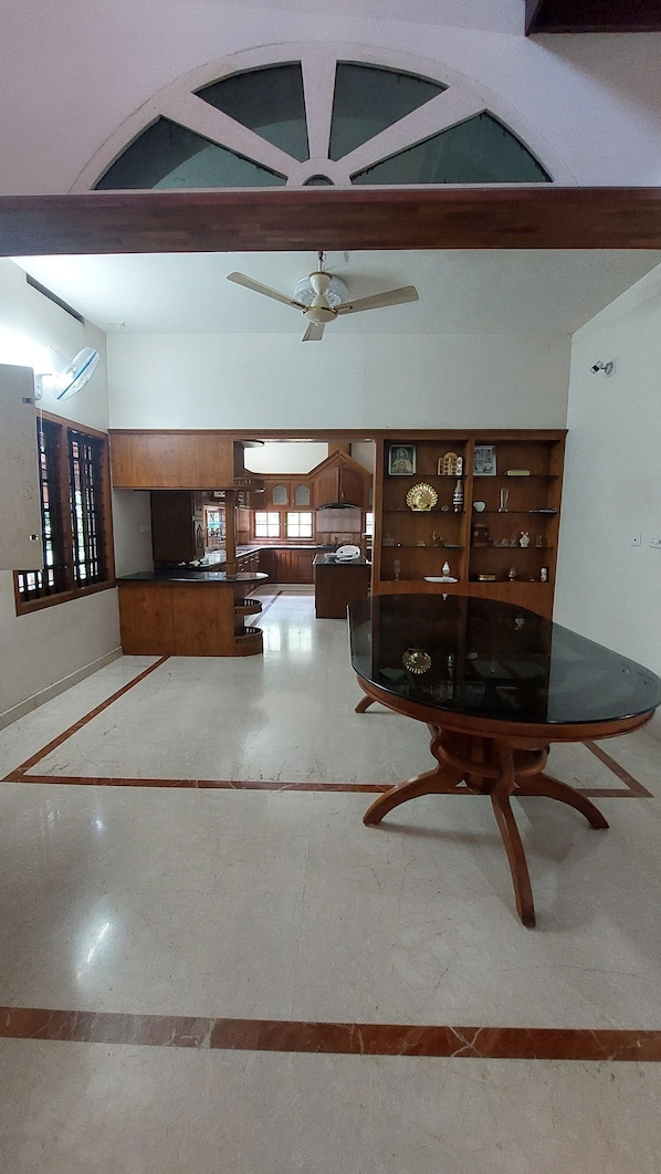 A dining area with a glass table and wooden chairs, a ceiling fan, and a view of the kitchen in the background.