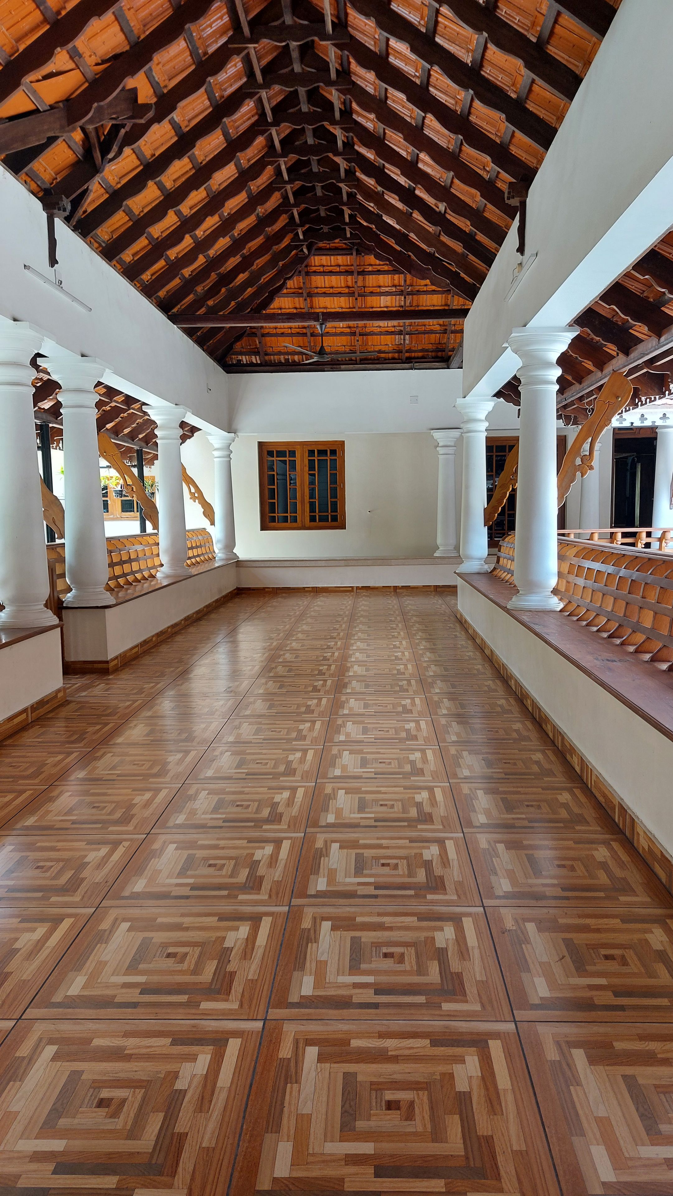 A spacious corridor with a patterned wooden floor, white columns, and a wooden ceiling.