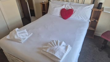 A neatly made bed with white linens, a red heart-shaped pillow, and folded towels on the bed.