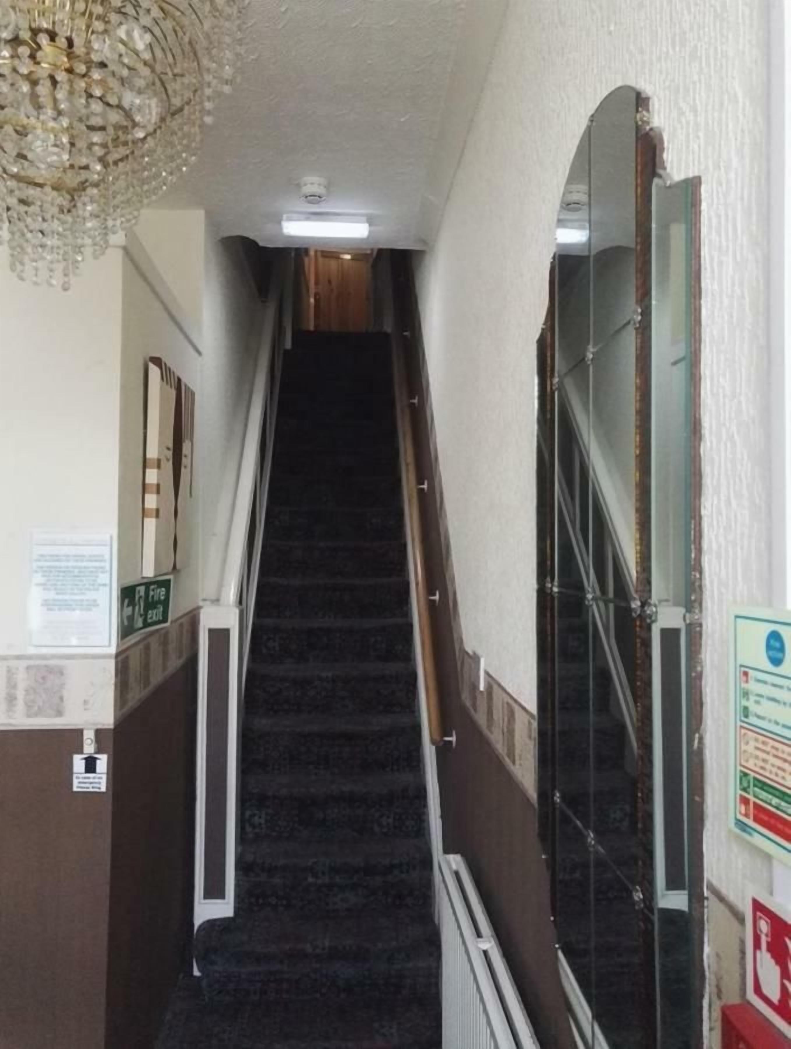 A narrow staircase with a chandelier, a fire exit sign, and a mirror on the wall.