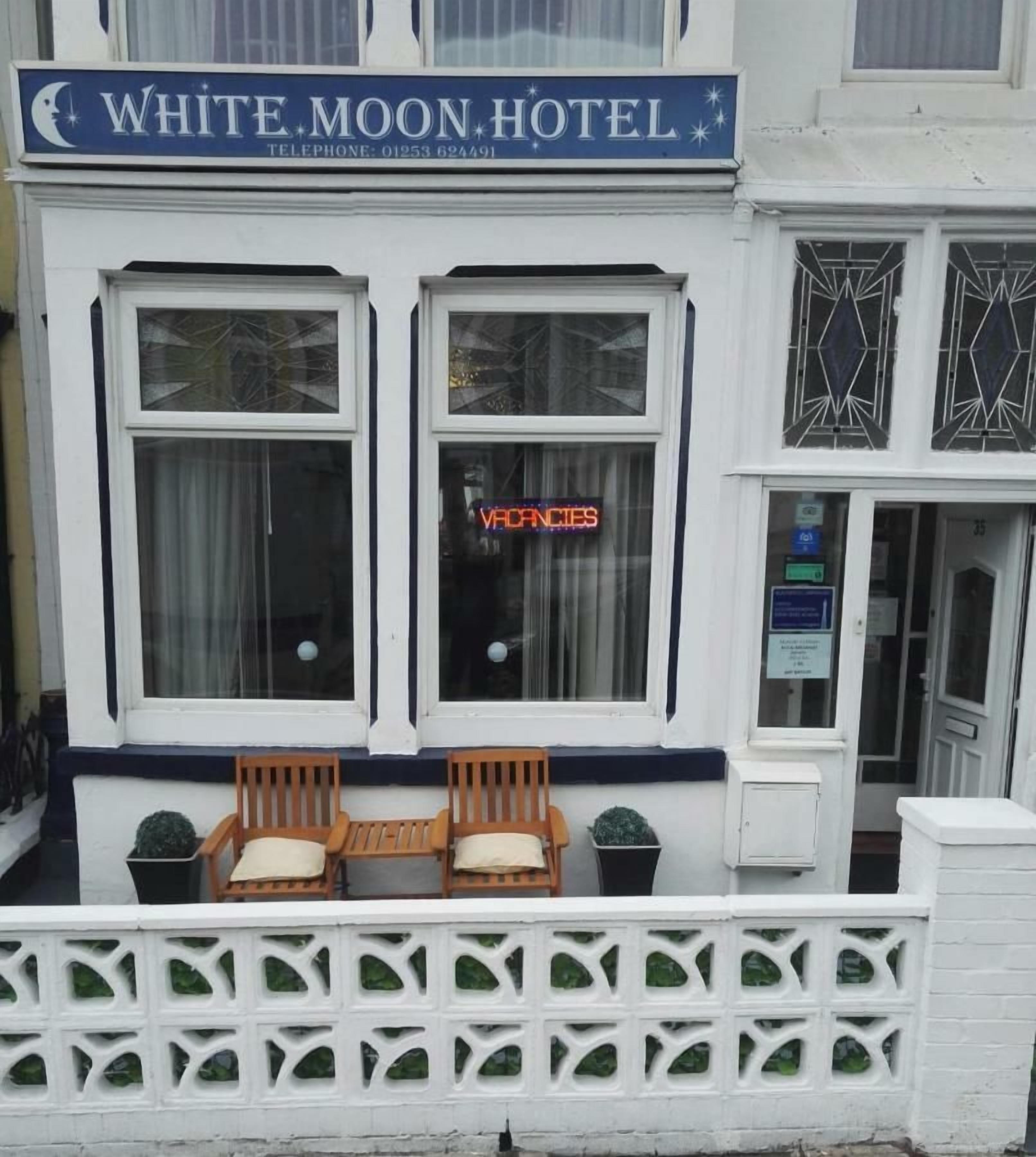 A hotel named White Moon Hotel with a signboard, two windows with curtains, and a white fence with greenery.
