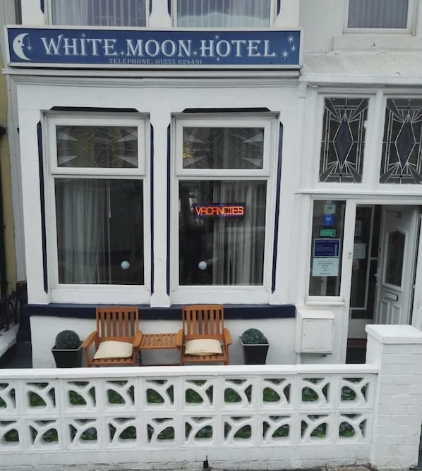 Un hotel llamado White Moon Hotel con un letrero, dos ventanas con cortinas y una cerca blanca con follaje.