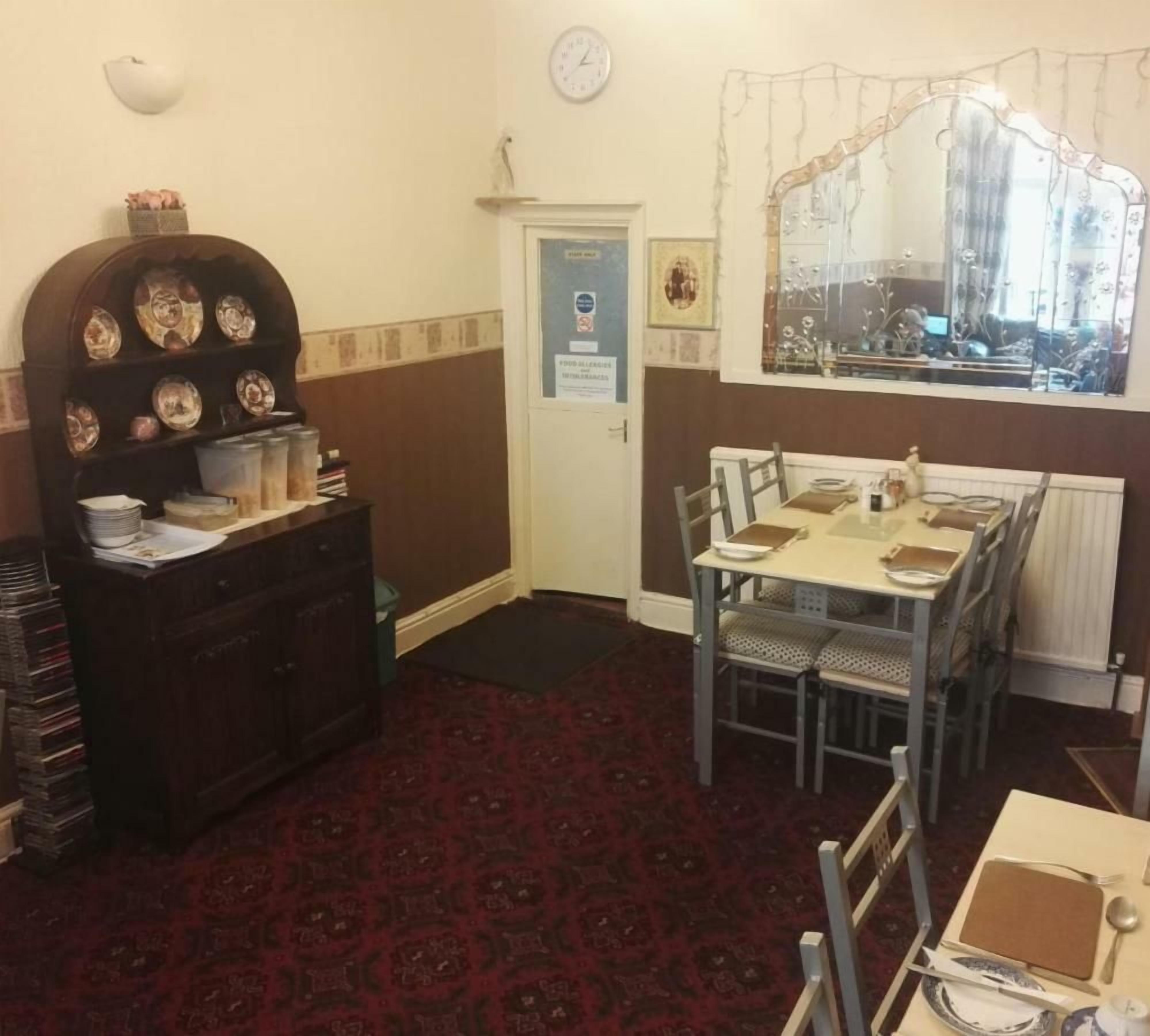 A dining area with a table set for four, a dark wooden cabinet with decorative plates, a clock on the wall, and a window with a floral curtain.