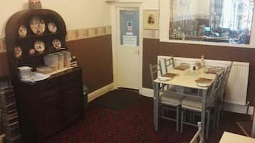 A dining area with a table set for four, a dark wooden cabinet with decorative plates, a clock on the wall, and a window with a floral curtain.