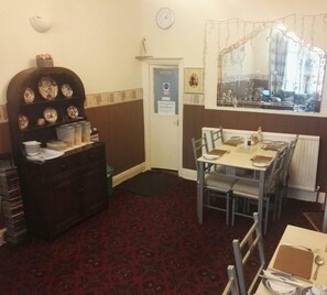 A dining area with a table set for four, a dark wooden cabinet with decorative plates, a clock on the wall, and a window with a floral curtain. - White Moon Hotel (Blackpool)