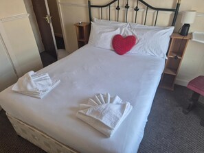 Una cama bien hecha con sábanas blancas, un cojín rojo con forma de corazón y toallas dobladas sobre la cama.