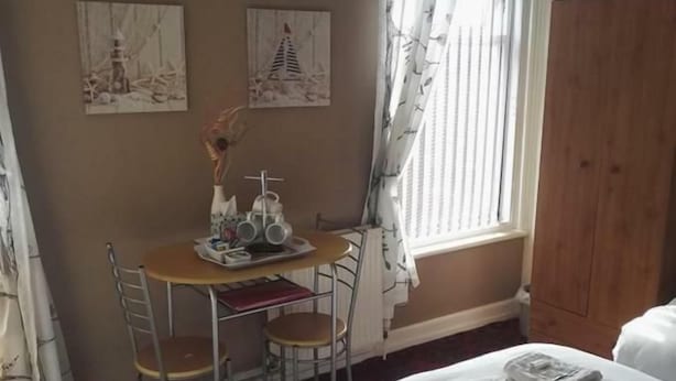 A hotel room with a bed, a desk with chairs, a window with curtains, and artwork on the wall. - White Moon Hotel (Blackpool)