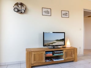 A wooden TV stand with a flat-screen TV displaying a beach scene, two framed pictures on the wall, and a small toy car decoration. - 9 in the Thomsen House (St. Peter-Ording)